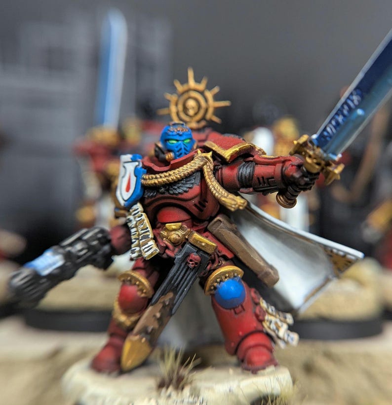 May include: A red and gold painted miniature figure of a space marine warrior holding a sword and a gun. The figure is wearing a white cape and has a blue face.