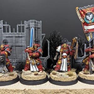 May include: Five red and gold painted miniature figures of space marines with swords and guns, standing on round bases. The figures are arranged in a line, with a banner behind them. The banner is red and white with a gold eagle emblem and the text "II".