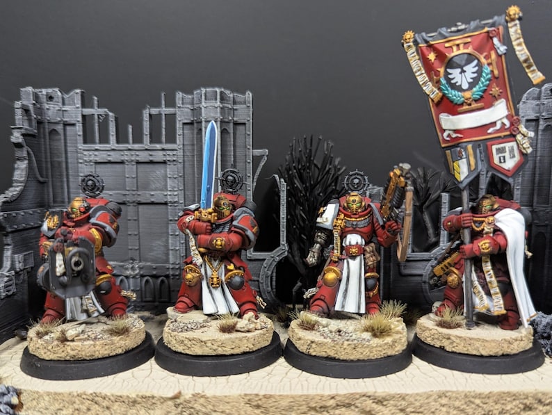 May include: Five red and gold painted miniature figures of space marines with swords and guns, standing on round bases. The figures are arranged in a line, with a banner behind them. The banner is red and white with a gold eagle emblem and the text "II".