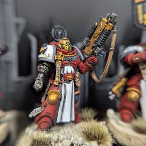 May include: A red and gold painted miniature of a space marine with a white tabard and a large gun. The miniature is posed in a dynamic stance, holding the gun in one hand and a sword in the other. The miniature is set on a base with a brown and green texture.