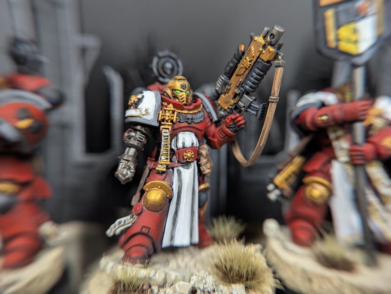 May include: A red and gold painted miniature of a space marine with a white tabard and a large gun. The miniature is posed in a dynamic stance, holding the gun in one hand and a sword in the other. The miniature is set on a base with a brown and green texture.