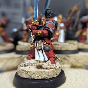 May include: A red and gold painted miniature figure of a space marine holding a blue sword. The figure is standing on a sandy base with a black base.