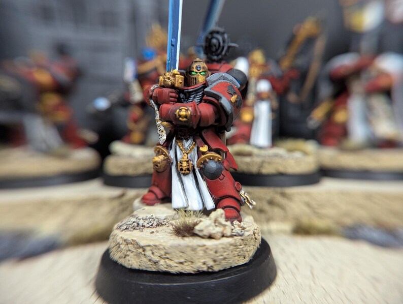 May include: A red and gold painted miniature figure of a space marine holding a blue sword. The figure is standing on a sandy base with a black base.