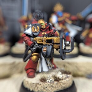 May include: A red and gold painted miniature figure of a space marine holding a large gun. The figure is standing on a round base with a brown and white textured surface.