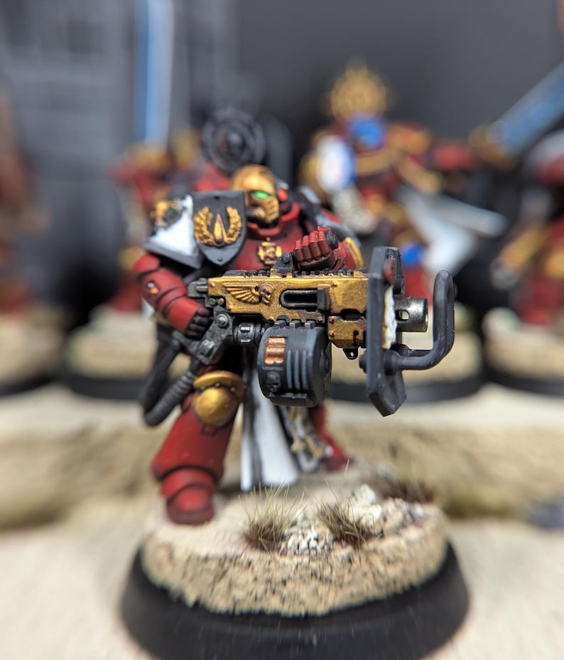 May include: A red and gold painted miniature figure of a space marine holding a large gun. The figure is standing on a round base with a brown and white textured surface.