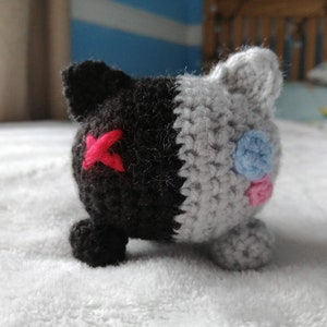 May include: A crocheted plush toy with a black and gray body. The toy has a red X on one side and blue and pink circles on the other side.