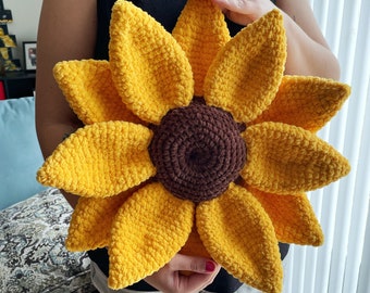 May include: A large crocheted sunflower pillow with a brown center and yellow petals. The pillow is being held by a person.