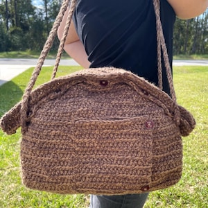 Puede incluir: Un bolso de crochet marrón con cierre de solapa y detalles de botones. El bolso se lleva al hombro y tiene una correa larga.