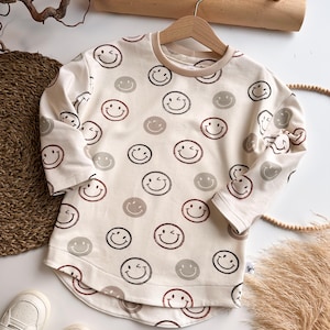 Könnte beinhalten: Ein cremefarbenes Langarmshirt mit abgerundetem Saum. Das Shirt hat ein Muster aus braunen, schwarzen und roten Smileys.