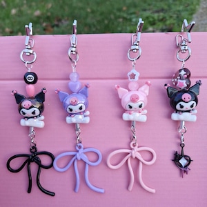 May include: Four keychains with cartoon characters in black, purple, and pink. Each keychain has a different charm, including a bow, star, and an 8-ball. The keychains are attached to silver-coloured clips and hang against a pink background.