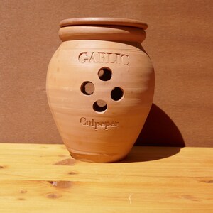 Puede incluir: Un tarro de ajo de terracota con las palabras "GARLIC" y "Culpeper" inscritas. El tarro tiene cuatro orificios de ventilación circulares y una tapa. Está sobre una superficie de madera con un fondo marrón.