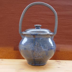 May include: A blue ceramic pot with a lid and a handle. The pot has a round body and a curved handle. The pot has a design on the front. The pot is sitting on a wooden surface.