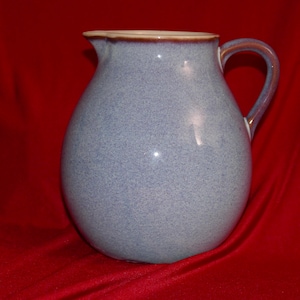 May include: A light blue ceramic pitcher with a speckled finish and a rounded body. The vessel features a curved handle on the right side and a smooth, light brown rim around the top opening, showcasing a simple, functional design for pouring liquids.