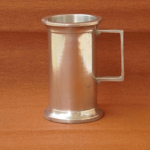 May include: A silver-coloured metal tankard with a rectangular handle. The tankard has a cylindrical body with a slightly flared base and a rim around the top. The tankard is made of a shiny metal and is set against a brown background.