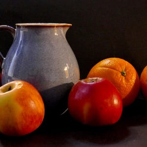 Könnte beinhalten: Ein Stillleben mit einer blauen Keramikkrug und Früchten. Es gibt rote und gelbe Äpfel und leuchtend orange Orangen. Der Hintergrund ist dunkel.