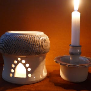 May include: A white ceramic oil warmer with a textured, grooved top and arched cutouts, accompanied by a white ceramic candle holder featuring a scalloped edge and a single lit white taper candle, creating a warm, glowing atmosphere.