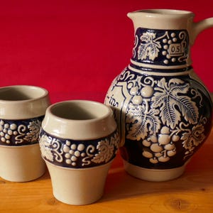Peut inclure: Une carafe en céramique et deux tasses assorties présentant un coloris bicolore beige et bleu cobalt. L'ensemble est orné de motifs en relief représentant des grappes de raisin et des feuilles de vigne. La carafe possède une anse et une marque 0,5L.