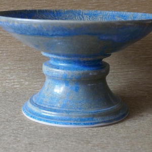 Op de afbeelding: Een keramische schaal op voet met een brede, ondiepe kom en een stevige, uitlopende basis. Het oppervlak is afgewerkt met een gespikkeld glazuur in tinten van levendig blauw en gedempt grijs, wat zorgt voor een textuur over de gehele buitenkant.