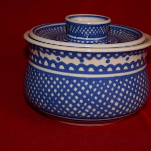 Peut inclure: Beurrier en céramique bleu et blanc avec couvercle. Le plat présente un motif répétitif de losanges bleus et une bordure festonnée blanche. Le couvercle a une petite poignée et un motif bleu et blanc assorti. Le plat est sur une surface rouge.