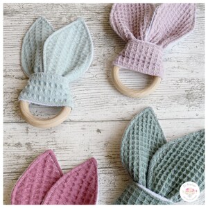 Puede incluir: Tres mordedores de madera con orejas de conejo de tela. Los mordedores son en tonos de verde, rosa y azul claro. La tela es una textura de tejido de waffle.