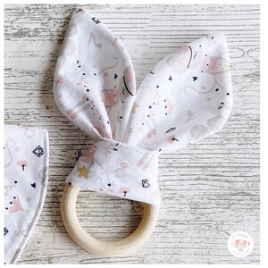 Puede incluir: Un juguete de dentición para bebés con un anillo de madera y orejas de conejo de tela. La tela blanca está estampada con unicornios rosas y grises, estrellas y flores. El anillo de madera es de color marrón claro.