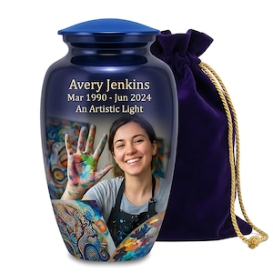 May include: A dark blue metal cremation urn featuring a custom photo of a person with paint-covered hands, the text Avery Jenkins, Mar 1990 - Jun 2024, and An Artistic Light. A matching dark purple velvet drawstring bag with gold cords sits beside it.
