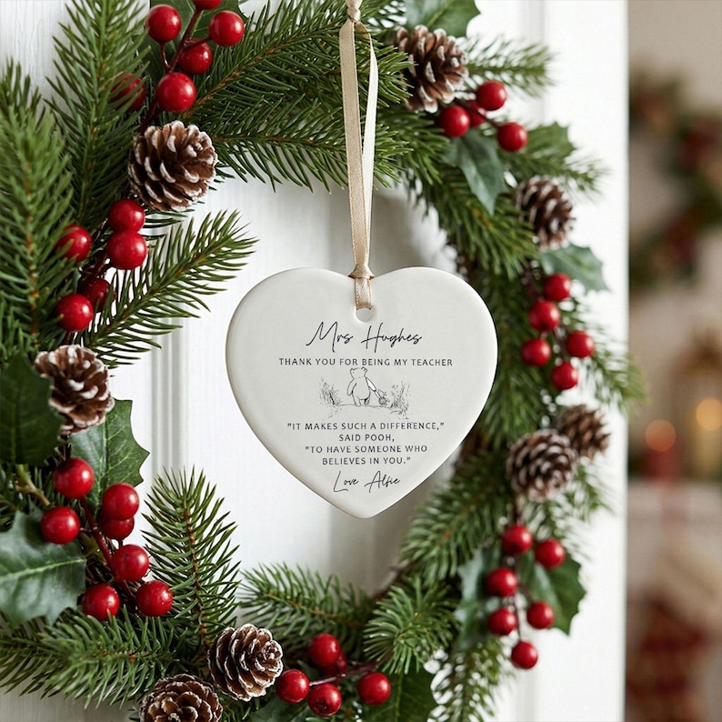 May include: A heart-shaped ceramic ornament with a beige ribbon, hanging in front of a Christmas wreath. The ornament has text that reads "Mrs. Hughes, Thank you for being my teacher." The wreath is made of green pine needles, red berries, and pine cones.