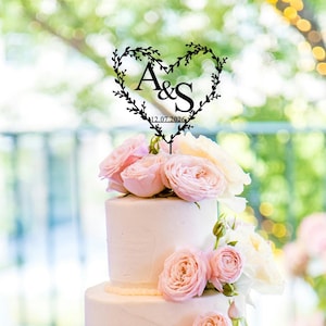 May include: A white wedding cake decorated with pink roses and a black heart-shaped topper with the initials "A&S" and the date "12.07.2026".