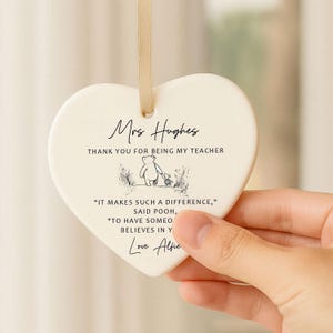 May include: A heart-shaped ceramic ornament with a cream finish and a gold ribbon for hanging. The ornament features the text "Mrs Hughes, Thank you for being my teacher" and a Winnie the Pooh illustration. Additional text includes a quote and a signature.