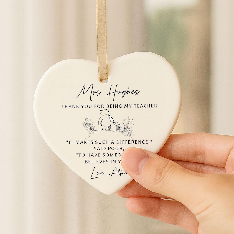 May include: A heart-shaped ceramic ornament with a cream finish and a gold ribbon for hanging. The ornament features the text "Mrs Hughes, Thank you for being my teacher" and a Winnie the Pooh illustration. Additional text includes a quote and a signature.