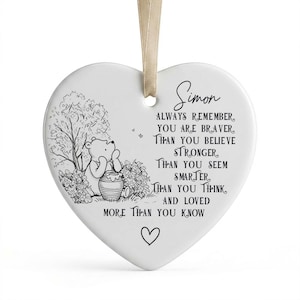 May include: A white heart-shaped ceramic ornament hanging from a beige ribbon. It features a black line drawing of Winnie the Pooh with a honey pot and the text: Simon, Always remember you are braver than you believe, stronger than you seem, smarter than you think, and loved more than you know, with a small heart icon below.
