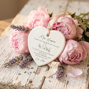 May include: A heart-shaped ceramic plaque with a beige ribbon, featuring text that reads "Mrs. Hughes, Thank you for being my teacher." The plaque is decorated with a Winnie the Pooh illustration and a quote. The plaque is surrounded by pink peonies and lavender.