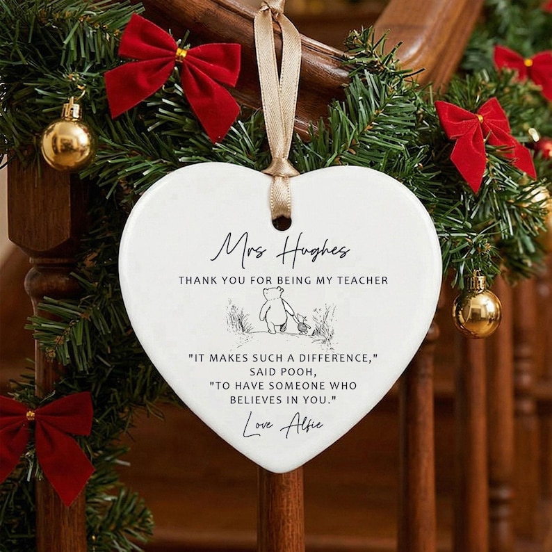 May include: A heart-shaped, white ceramic ornament with a gold ribbon for hanging. The ornament has text that reads "Mrs Hughes, Thank you for being my teacher" and a quote from Winnie the Pooh. The ornament is decorated with a Winnie the Pooh illustration and the name "Love Alfie".