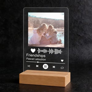 May include: A clear acrylic night light with a photo of three people hugging. The light has a wooden base and displays the text "Friendships" and the artist's name. The design resembles a music player interface.