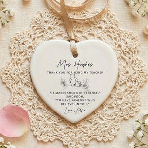 May include: A white heart-shaped ceramic ornament with a beige ribbon for hanging. The ornament has text that reads "Mrs Hughes, THANK YOU FOR BEING MY TEACHER" and a quote from Winnie the Pooh. The ornament is on a beige lace doily.