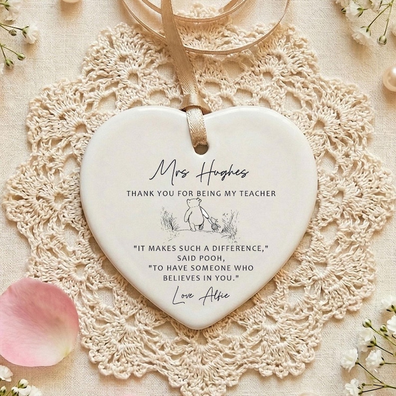 May include: A white heart-shaped ceramic ornament with a beige ribbon for hanging. The ornament has text that reads "Mrs Hughes, THANK YOU FOR BEING MY TEACHER" and a quote from Winnie the Pooh. The ornament is on a beige lace doily.