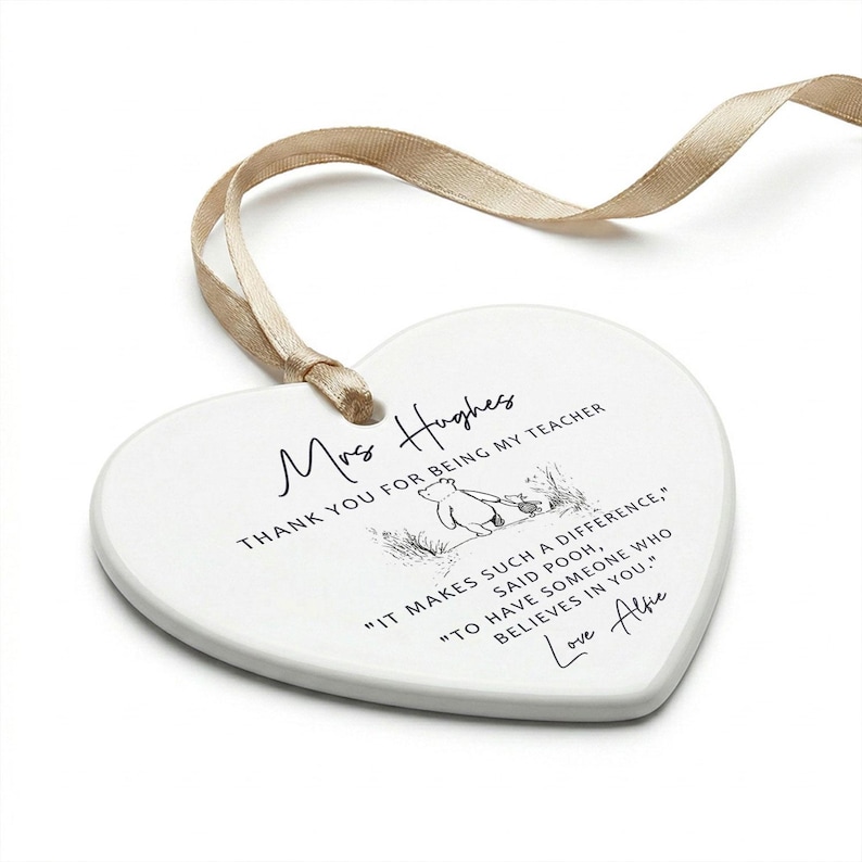 May include: A white heart-shaped ceramic ornament with a beige ribbon. The ornament has text that reads "Mrs Hughes, THANK YOU FOR BEING MY TEACHER" and a Winnie the Pooh illustration. The quote reads "IT MAKES SUCH A DIFFERENCE, SAID POOH, TO HAVE SOMEONE WHO BELIEVES IN YOU." Signed "Love Alfie."