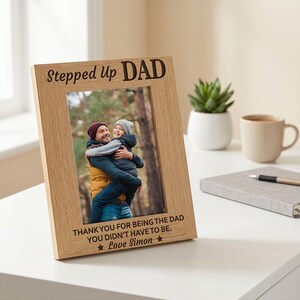 May include: A light oak picture frame with the words "Stepped Up DAD" at the top and a photo of a father and child. The frame includes the text "THANK YOU FOR BEING THE DAD YOU DIDN'T HAVE TO BE. Love Simon."