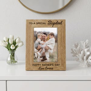 May include: A light brown wooden photo frame with the text "TO A SPECIAL Stepdad" and "HAPPY FATHER'S DAY Love Emma". The frame contains a photograph of a man holding two children. The frame is displayed on a white surface.