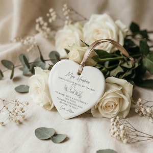 May include: A heart-shaped ceramic ornament with a gold ribbon, featuring text that reads "Mrs. Hughes, Thank you for being my teacher." The ornament is surrounded by white roses and greenery. The text also includes a quote from Winnie the Pooh.