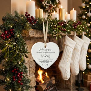 May include: A heart-shaped ceramic ornament with a cream-colored finish, hanging from a beige ribbon. The ornament has text that reads "Mrs. Hughes, Thank you for being my teacher." The background includes a fireplace, stockings, and Christmas decorations.