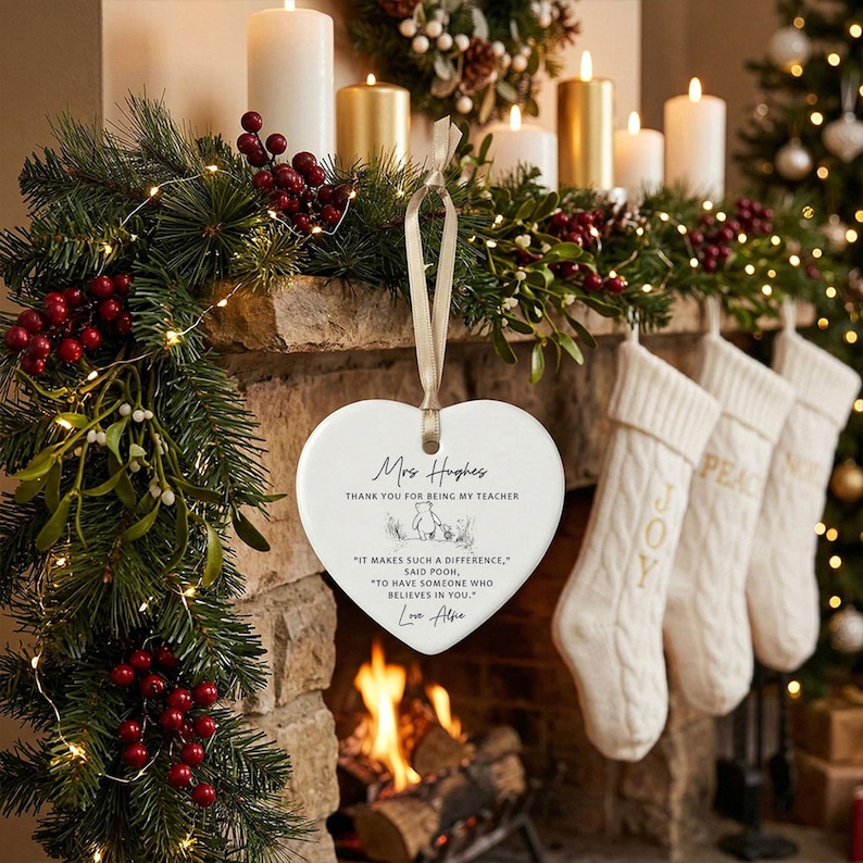 May include: A heart-shaped ceramic ornament with a cream-colored finish, hanging from a beige ribbon. The ornament has text that reads "Mrs. Hughes, Thank you for being my teacher." The background includes a fireplace, stockings, and Christmas decorations.