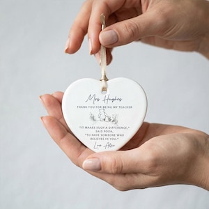 May include: A white heart-shaped ceramic ornament with a beige ribbon for hanging. The ornament has black text that reads "Mrs Hughes, THANK YOU FOR BEING MY TEACHER." Below is a Winnie the Pooh illustration and a quote.
