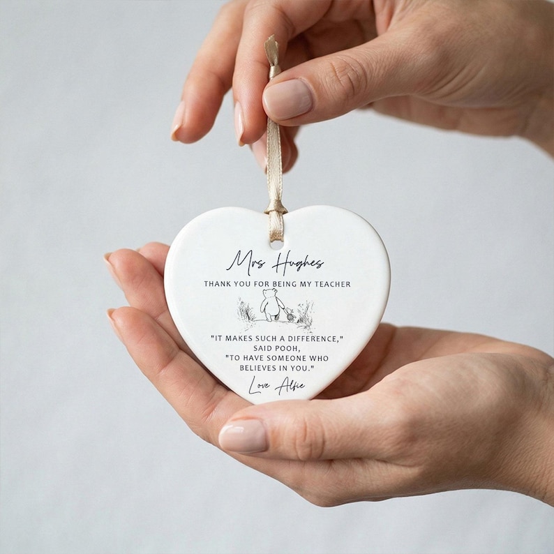 May include: A white heart-shaped ceramic ornament with a beige ribbon for hanging. The ornament has black text that reads "Mrs Hughes, THANK YOU FOR BEING MY TEACHER." Below is a Winnie the Pooh illustration and a quote.