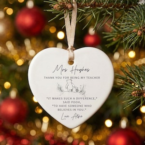 May include: A white heart-shaped Christmas ornament with a beige ribbon. The ornament has the text "Mrs Hughes, Thank you for being my teacher" and a quote from Winnie the Pooh. The ornament is hanging on a Christmas tree.
