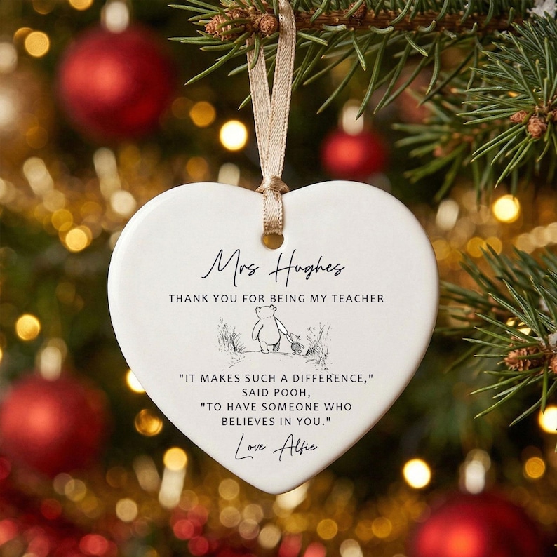 May include: A white heart-shaped Christmas ornament with a beige ribbon. The ornament has the text "Mrs Hughes, Thank you for being my teacher" and a quote from Winnie the Pooh. The ornament is hanging on a Christmas tree.