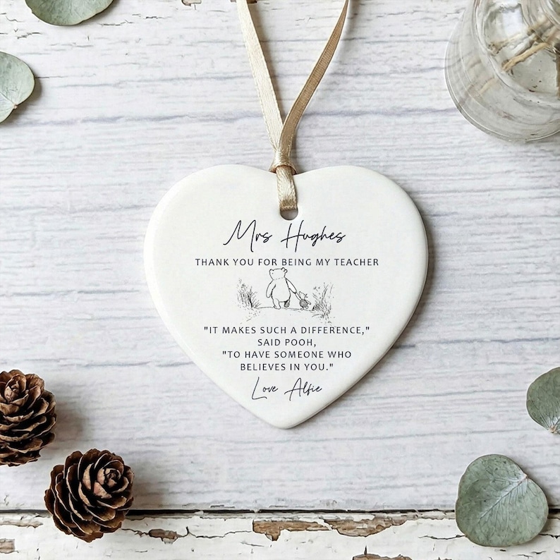 May include: A heart-shaped ceramic ornament with a cream-colored finish. The ornament has a beige ribbon for hanging and features the text "Mrs Hughes, Thank you for being my teacher." Below is a Winnie the Pooh illustration and a quote.
