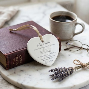 May include: A white heart-shaped ceramic ornament with a gold ribbon, inscribed with a message for a teacher. The ornament rests on a book next to a cup of coffee, glasses, and a bundle of lavender.