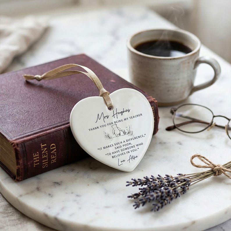 May include: A white heart-shaped ceramic ornament with a gold ribbon, inscribed with a message for a teacher. The ornament rests on a book next to a cup of coffee, glasses, and a bundle of lavender.