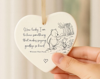 May include: Cream-coloured heart-shaped ceramic ornament featuring a Winnie the Pooh illustration and the quote "How lucky I am to have something that makes saying goodbye so hard." A beige ribbon is attached for hanging.